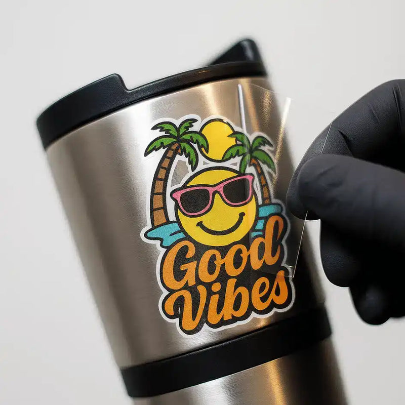 A stainless steel travel mug with a ’Good Vibes’ sticker being applied.